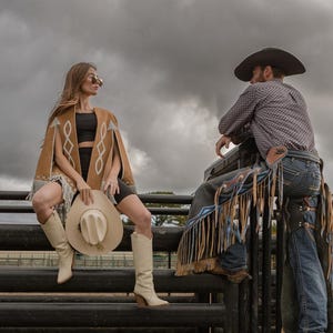May include: A woman wearing a tan suede cape with fringe and a cowboy hat, paired with cream-colored boots. A man in a cowboy hat and chaps is also present. The scene is set outdoors with a cloudy sky.