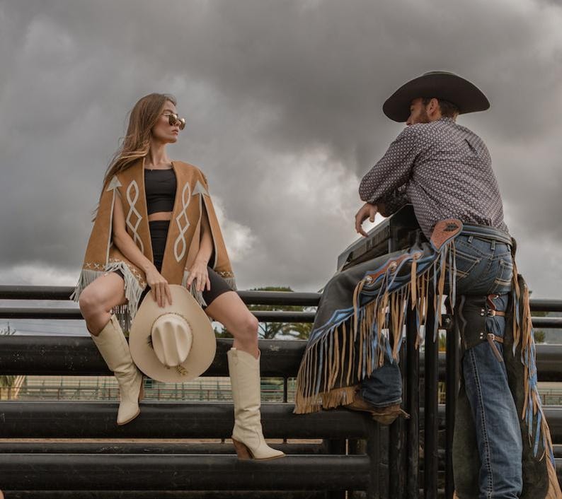 May include: A woman wearing a tan suede cape with fringe and a cowboy hat, paired with cream-colored boots. A man in a cowboy hat and chaps is also present. The scene is set outdoors with a cloudy sky.
