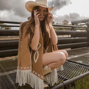 May include: A tan cowboy hat with a decorative band and sunglasses. A brown suede poncho with white geometric designs and fringe. The poncho is paired with white cowboy boots. The background features a cloudy sky and a metal structure.