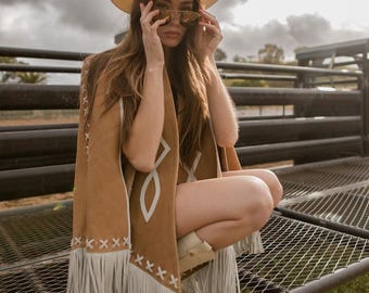May include: A tan cowboy hat with a decorative band and sunglasses. A brown suede poncho with white geometric designs and fringe. The poncho is paired with white cowboy boots. The background features a cloudy sky and a metal structure.