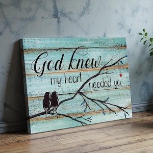 May include: A wooden wall art piece with a distressed, light blue background. The artwork features a black script that reads "God knew my heart needed you." Two black birds perch on a branch with a small red heart. The overall theme is love and faith.