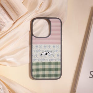 May include: A phone case with a Snoopy design. The case features a gray border and a three-section design: pink stripes, a floral pattern, and green plaid. Snoopy is depicted sleeping in the center of the case.