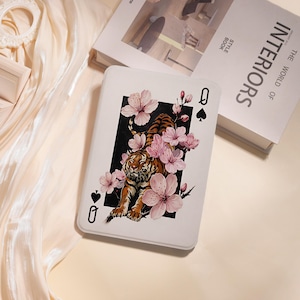 May include: A rectangular white card featuring a central illustration of a tiger surrounded by pink cherry blossoms set against a black rectangular background. The card displays the letter Q and a black spade symbol in the top right and bottom left corners.