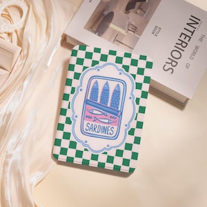 Op de afbeelding: Een rechthoekige, bedrukte hoes met een groen en wit geblokt patroon. De hoes heeft een witte rand met een cartoon illustratie van sardines in een blik, met het woord "SARDINES" eronder. De hoes ligt op een crèmekleurig oppervlak.