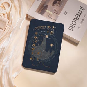 Può includere: Una custodia per tablet blu scuro con un'illustrazione dorata del Castello di Hogwarts e il testo "Hogwarts School of Witchcraft & Wizardry". Il design include impronte e stelle. Un libro intitolato "Interiors" è sullo sfondo.