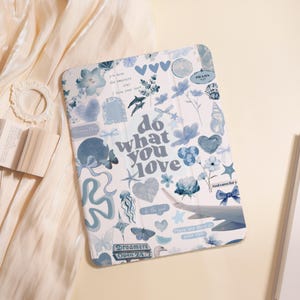 May include: A white tablet case adorned with blue illustrations of flowers, hearts, and butterflies, alongside the words "do what you love." The case also features the text "Dreamers Open 24/7" and "There are stars in your eyes."