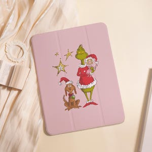 May include: A light pink tablet case featuring a printed illustration of a green creature in a red Santa suit holding a small character, accompanied by a brown dog wearing a red Santa hat and a green collar, alongside three gold stars.