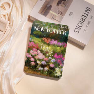 May include: A copy of The New Yorker magazine from July 19, 1982, featuring a colorful floral painting on the cover. The magazine is placed on a cream-colored surface next to a book titled "Interiors".