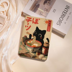 May include: A beige-colored notebook featuring a black cat eating ramen with chopsticks. The illustration has Japanese text and a red sun. The notebook is surrounded by other books.