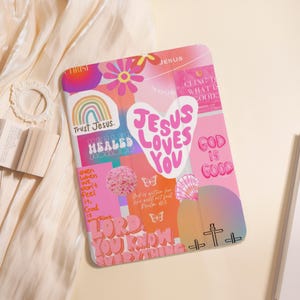 Peut inclure: Une coque de tablette rose et blanche avec un motif de collage. Le design comprend un cœur avec le texte "Jesus Loves You", un arc-en-ciel, des fleurs et d'autres phrases inspirantes. La coque présente une croix et les mots "God is Good".