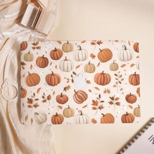 Pode incluir: Uma capa bege para laptop com um padrão repetido de abóboras em tons de laranja, branco e bege, juntamente com folhas marrons. A capa tem um recorte para o logotipo da Apple. O tema é outono.