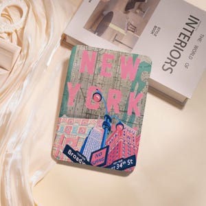 May include: A rectangular case with a map of New York City, the city name in pink block letters. The case has illustrations of buildings, street signs, and a blue and pink tower. The case is on a cream-coloured surface.