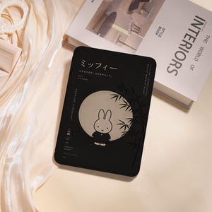 Könnte beinhalten: Eine schwarze, rechteckige E-Reader-Hülle mit einer Grafik des Hasen Miffy vor einem großen, blassen Mond. Das Design enthält japanischen Text, Bambuszweige auf der rechten Seite und kleine Symbole. Die Oberfläche ist matt und mit weißem Text bedruckt.