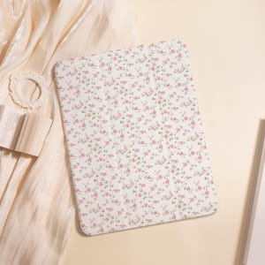 Może przedstawiać: Białe etui na tablet z kwiatowym wzorem małych różowych róż i zielonych liści. Etui jest prostokątne z zaokrąglonymi rogami. Tło obejmuje kremową tkaninę i małą otwartą książkę.