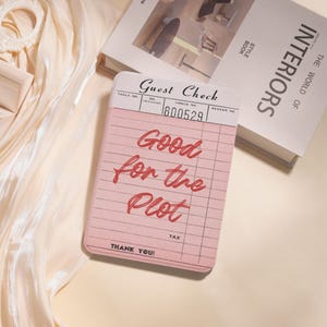 May include: A pink guest check notepad with the words "Good for the Plot" in red script. The notepad has a lined design and the words "Guest Check" at the top. A book titled "INTERIORS" is in the background.