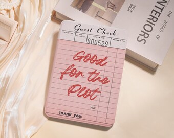 May include: A pink guest check notepad with the words "Good for the Plot" in red script. The notepad has a lined design and the words "Guest Check" at the top. A book titled "INTERIORS" is in the background.