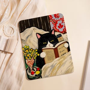 May include: A rectangular tablet case featuring a black and white cat reading a book titled El médico by Noah Gordon. The illustration includes a vase of yellow flowers, a small table with red and yellow fruit, and a red floral pattern background.