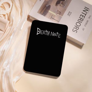 May include: A black notebook with the words "Death Note" in white script. The notebook is rectangular with rounded corners. The image is on a cream-colored surface, with a book titled "Interiors" in the background.