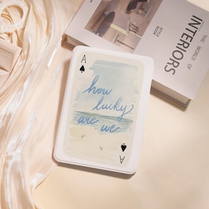 May include: A playing card with the Ace of Spades, displaying the text "how lucky are we" in blue script. The card's design includes a watercolor beach scene with birds. The card is set against a soft, neutral background.