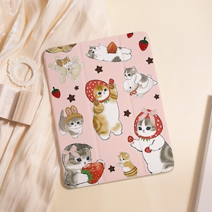 Op de afbeelding: Roze tablethoes met cartoonkatjes, aardbeien en sterrenmotieven. De hoes heeft een opvouwbare hoes en een speels ontwerp. De katjes staan in verschillende poses, sommigen dragen aardbeienhoeden, anderen houden aardbeien vast.