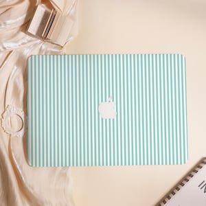 May include: A laptop case with a vertical striped pattern in light teal and white. The Apple logo is visible in white. The case is on a cream-colored surface with a book and decorative frame.