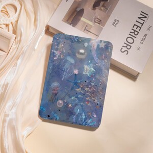 May include: A blue tablet case with a shimmering underwater design featuring jellyfish, starfish, and bubbles. The case has a glossy finish and is decorated with glitter and small details. The design evokes a dreamy, aquatic theme.