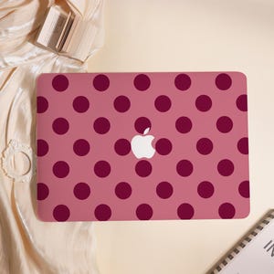 May include: A pink laptop case featuring a pattern of large, dark red polka dots. The Apple logo is in the centre. The case is on a cream-coloured surface with a small book and decorative frame.