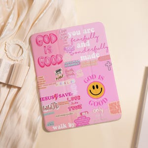 May include: Pink tablet case with various text stickers, including "God is Good," a smiley face, and religious phrases. The case has a soft, matte finish and a rectangular shape. The overall theme is inspirational and faith-based.