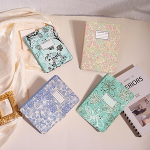 May include: Four rectangular composition books with distinct floral patterns. One is light blue with black floral swirls, one is pink and yellow with small flowers, one is blue with tiny blossoms, and one is teal with white and gold floral outlines.