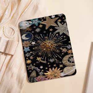 Op de afbeelding: Een zwarte decoratieve mat met een geborduurd hemels ontwerp. De mat heeft gouden, zilveren en blauwe sterren, planeten en een zon tegen een zwarte achtergrond. Het ontwerp bevat sterren in verschillende maten en vormen.