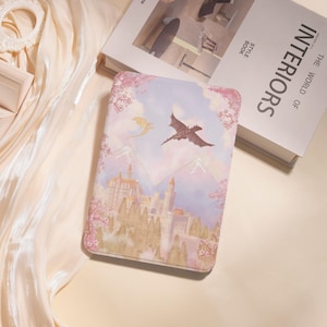 May include: A rectangular case featuring a fantasy landscape with a castle, mountains, and flying dragons. The case has a pastel color palette with pink cherry blossoms around the edges. A book titled "INTERIORS" is in the background.