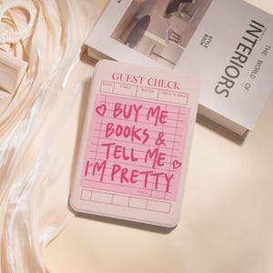 May include: A light pink guest check with the text "BUY ME BOOKS & TELL ME I'M PRETTY" in pink. The check is on a cream-colored surface, with a book titled "INTERIORS" in the background.