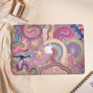 Op de afbeelding: Een rechthoekige harde laptophoes met een levendig, abstract geschilderd ontwerp met wervelende penseelstreken in roze, paarse, blauwe en gele tinten, gecentreerd rond een witte uitsparing in de vorm van het Apple-logo.