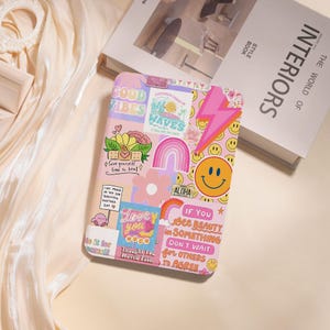 May include: A pink phone case covered in colorful stickers with phrases like "Good Vibes," "Waves," and "Love You Much." The case features a smiley face, a rainbow, and a pink lightning bolt design. The case is rectangular.