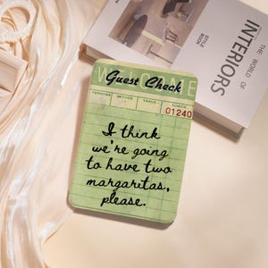 May include: A green guest check with the words "Guest Check" at the top. The check reads "I think we're going to have two margaritas, please." A book titled "INTERIORS" is in the background.