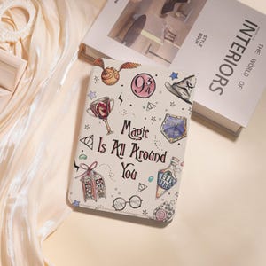 Peut inclure: Une housse de tablette blanche avec un design coloré représentant un Vif d'or, une coupe de maison Poudlard, et le texte "Magic Is All Around You". D'autres illustrations incluent un chapeau de sorcier, une potion et des lunettes.