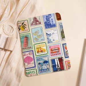 May include: A cream-colored travel-themed notebook featuring postage stamp designs with illustrations of the Eiffel Tower, a red double-decker bus, and the Leaning Tower of Pisa. The stamps include text such as "ITALY", "LONDON", and "MILANO".