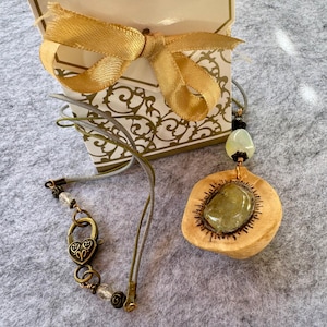 May include: A necklace with a wooden pendant featuring a green gemstone. The pendant is surrounded by a carved sun design. The necklace has a heart-shaped clasp and is made with brown leather cord and clear beads.