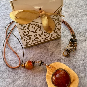 May include: A brown leather necklace with a wooden pendant featuring a round amber stone. The pendant is attached to a smaller amber bead and a silver clasp. The necklace is packaged in a white gift box with a gold ribbon.