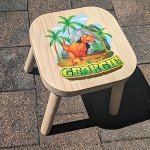May include: A wooden stool with a dinosaur and jungle scene design. The stool is painted in natural wood tones and features the name "George" in green letters.