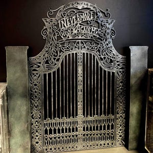 May include: A decorative metal gate with the words "Nevermore Academy" at the top. The gate is silver with intricate scrollwork and vertical bars. Two textured pillars flank the gate.