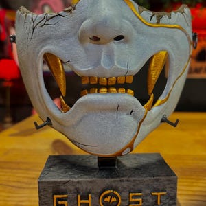 May include: A detailed, light blue and gold mask with a menacing design, featuring sharp teeth and a cracked texture. The mask is mounted on a dark gray base with the text "GHOST OF YOTE!" in gold lettering. The mask is a decorative item.