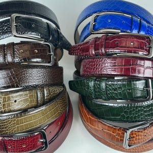 Può includere: Pile di cinture in pelle di vari colori, tra cui nero, marrone, blu, bordeaux, verde e beige. Ogni cintura presenta una fibbia color argento e un motivo strutturato a forma di coccodrillo. Le cinture sono disposte in due pile separate su sfondo bianco.