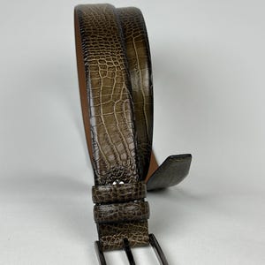 Peut inclure: Une ceinture en cuir marron avec un motif textur&eacute; en forme de crocodile. La ceinture est dot&eacute;e d'une boucle de couleur argent et d'une boucle assortie. La ceinture est enroul&eacute;e, mettant en valeur son design et sa texture.