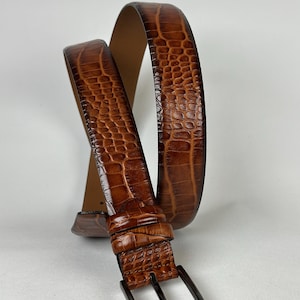 Peut inclure: Une ceinture en cuir marron avec un motif textur&eacute; en forme de crocodile. La ceinture est dot&eacute;e d'une boucle rectangulaire en m&eacute;tal fonc&eacute; et d'un int&eacute;rieur marron clair. La ceinture est incurv&eacute;e, mettant en valeur son design et sa texture.
