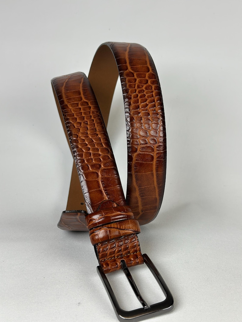Peut inclure: Une ceinture en cuir marron avec un motif textur&eacute; en forme de crocodile. La ceinture est dot&eacute;e d'une boucle rectangulaire en m&eacute;tal fonc&eacute; et d'un int&eacute;rieur marron clair. La ceinture est incurv&eacute;e, mettant en valeur son design et sa texture.