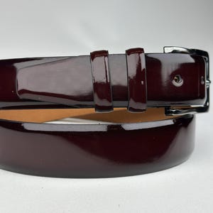 Pode incluir: Um cinto de couro bord&ocirc; com acabamento brilhante e fivela prateada.