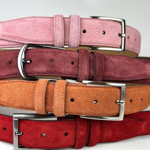 Könnte beinhalten: Ein Stapel aus vier Wildledergürteln in den Farben Hellrosa, Dunkelrosa, Orange und Rot. Jeder Gürtel verfügt über eine silberfarbene Metallschnalle, eine passende Lederschlaufe und vorgestanzte Löcher entlang des Riemens zur Größenanpassung.