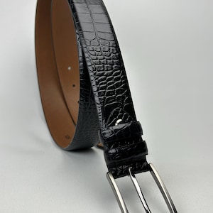 Peut inclure: Une ceinture noire avec un motif textur&eacute; en forme de peau de crocodile. La boucle est de couleur argent&eacute;e et l'int&eacute;rieur est marron. La ceinture est incurv&eacute;e, montrant sa flexibilit&eacute; et son design. Accessoire de mode.