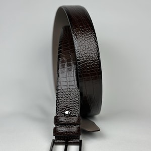 Peut inclure: Une ceinture marron fonc&eacute; avec un motif textur&eacute; en forme de crocodile. La ceinture a une boucle rectangulaire de couleur argent&eacute;e et une boucle de ceinture assortie. La ceinture en cuir est enroul&eacute;e, mettant en valeur son design et sa texture.